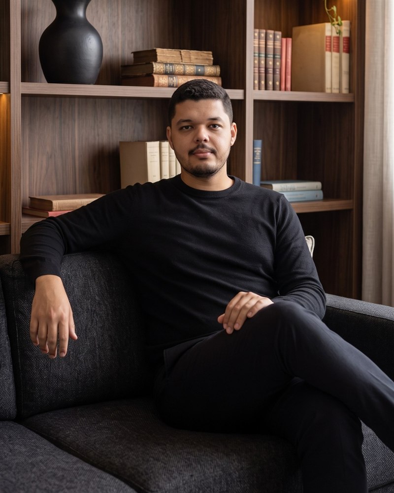 Ramon Alves, psicólogo, sentado em um sofá diante de uma estante de livros